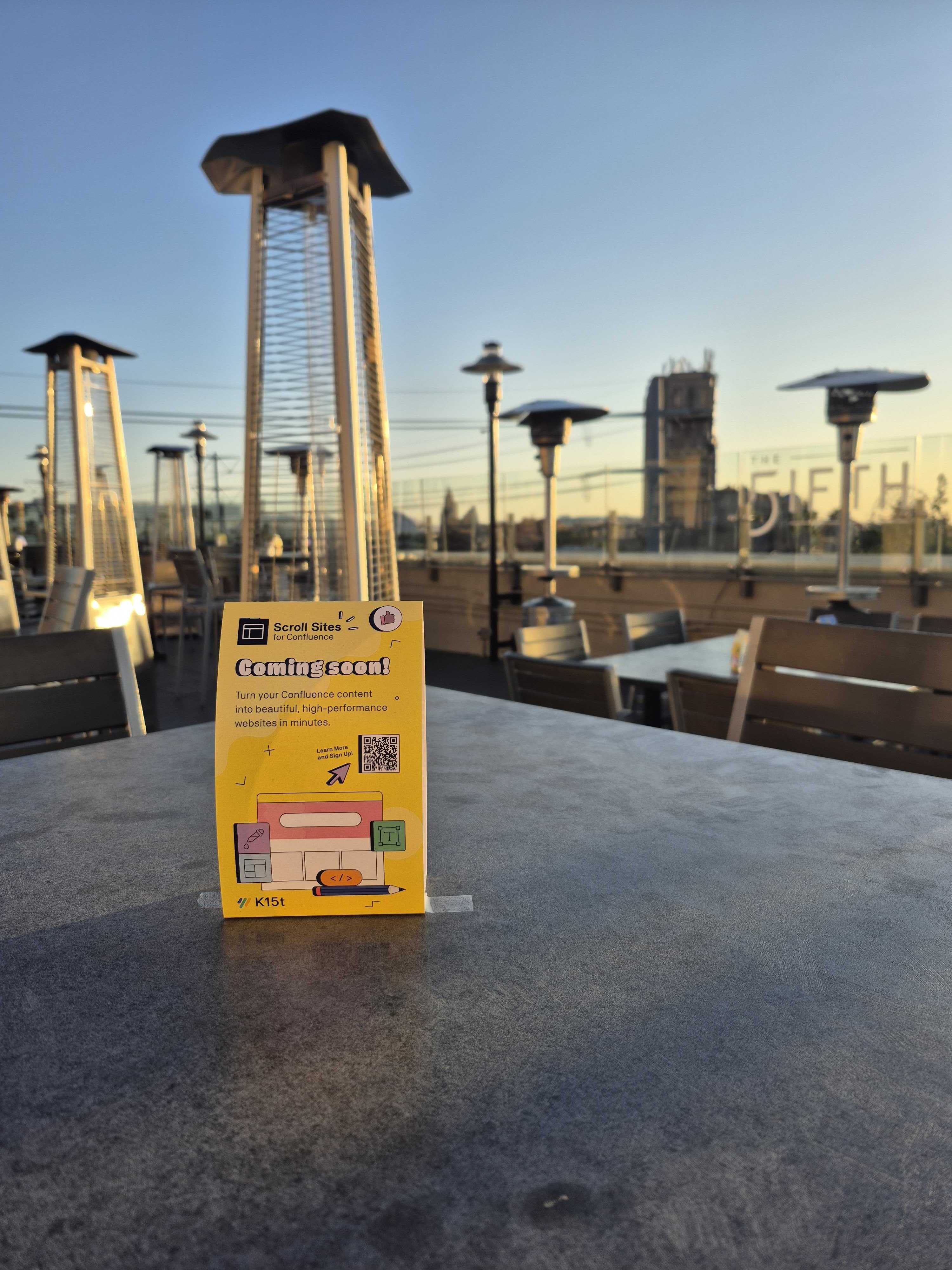 1000154570.jpgYellow event signage on a table at an outdoor rooftop bar with tall patio heaters under a clear sky.