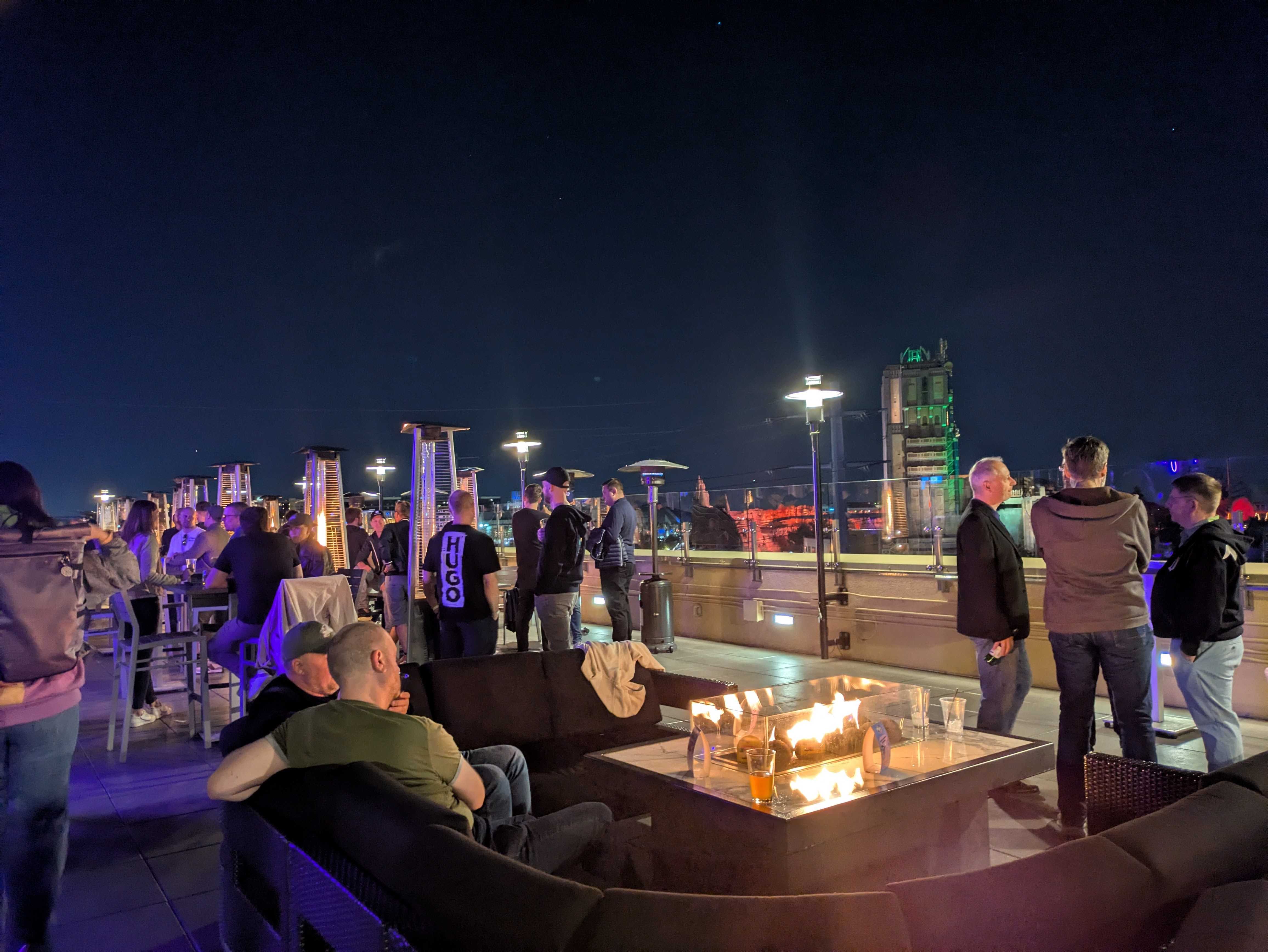 A night scene of the K15t Hocketse rooftop event with people gathering around a fire pit and patio heaters under the Anaheim night sky.